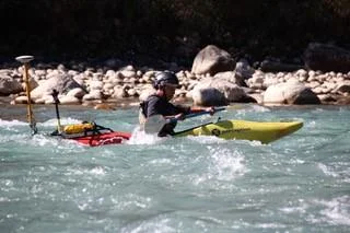Surveyor paddling a kayak with a GNSS antenna over a shallow gravel river