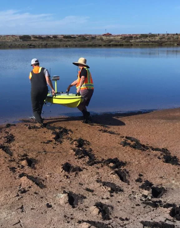 Two people carrying the CEE-USV across soft mud to the water