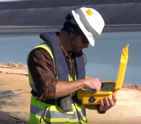 NewFields surveyor in hard hat and hi-vis using a yellow controller case beside a tailings pond