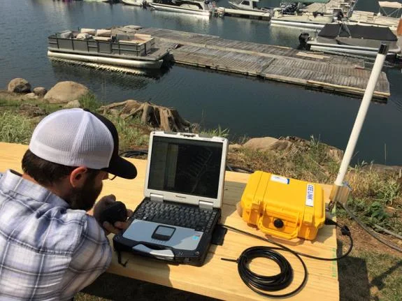Operator at a laptop beside a yellow CEE LINK base station on a dock with moored boats behind